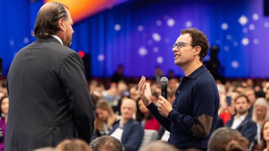 Dario Amodei, with curly hair and glasses and wearing a navy cardigan with brown elbow patches, holds a microphone and gestures as he talks to Marc Benioff, seen from behind in a dark pinstripe suit, surrounded by a seated audience in a hall lit in blue and orange