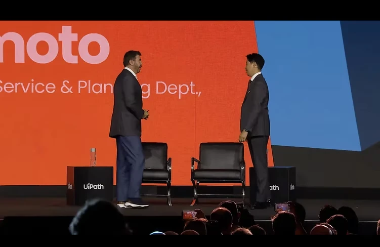 Daniel Dines and Kei Yamamoto standing on stage beside two black armchairs, in front of an orange screen.